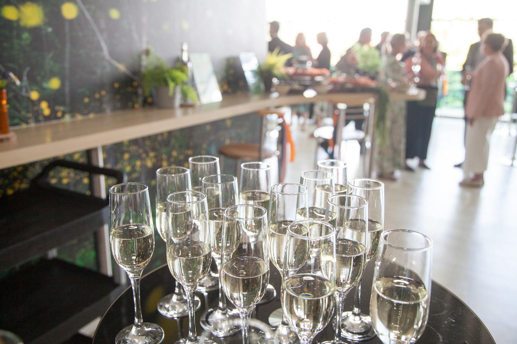A table with numerous champagne flutes filled with bubbly, set against a vibrant floral backdrop. In the blurred background, people are mingling at a social event.